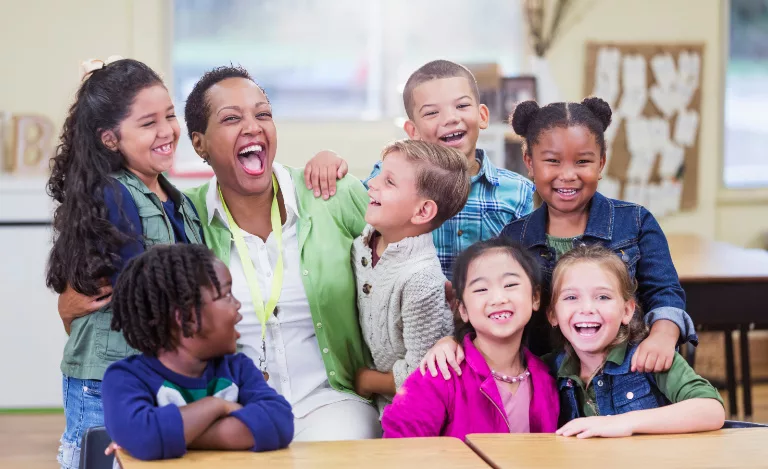 A teacher and seven young students smile and laugh while posing together in a classroom.