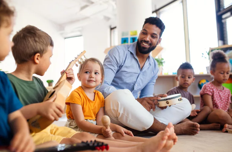 A group of young children sit on the floor with musical instruments, including a tambourine, a guitar, and a shaker, while an adult sits with them, smiling and interacting.