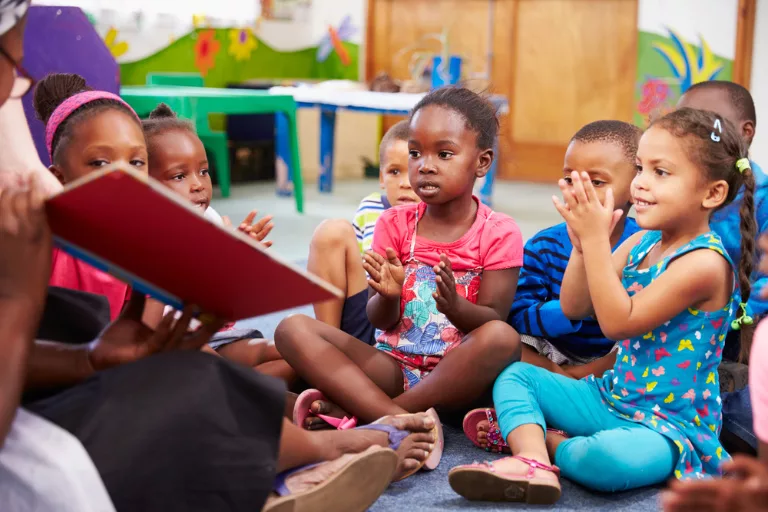 A group of children sit on the floor, clapping and listening to an adult reading from a book in a colorful classroom.