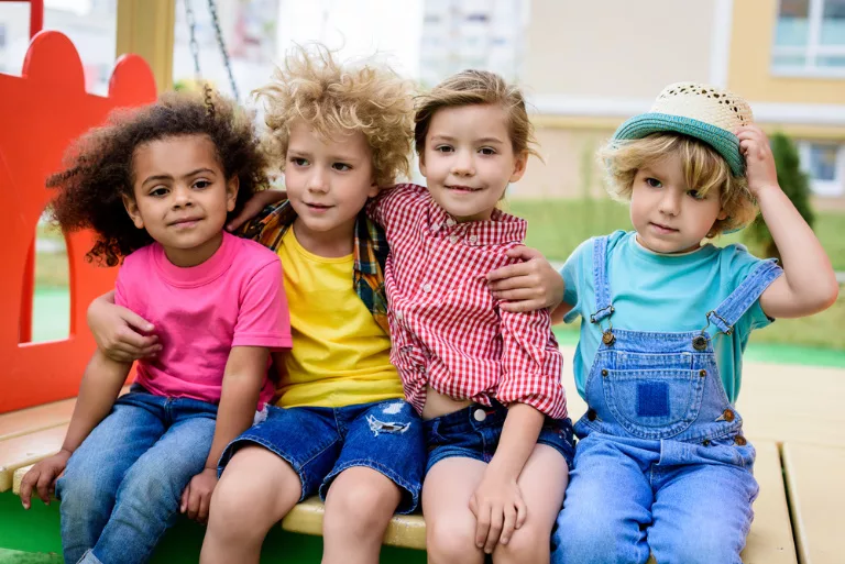 Four children wearing colorful clothes sit closely together on a play structure outdoors, smiling at the camera.